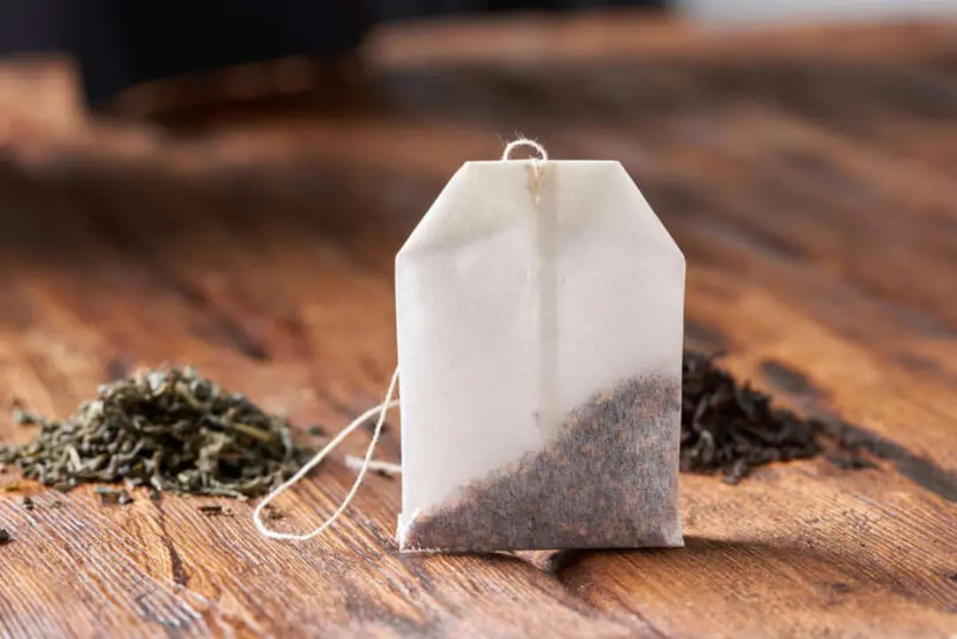 کیسه چای کیسه‌ای (Tea bag) در کنار برگ‌های خشک چای روی میز چوبی